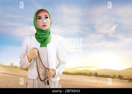 Giovani asiatici donna musulmana in velo in piedi e pregando con grani di preghiera sulla sabbia con cielo blu sullo sfondo Foto Stock