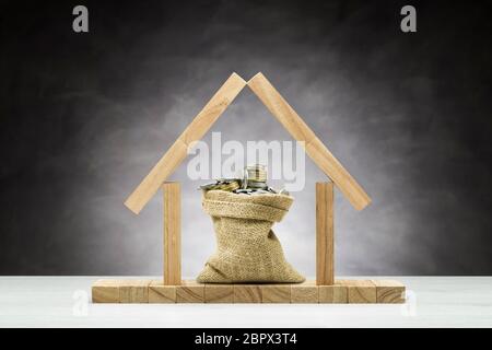 Concetto di protezione, casa in legno su uno sfondo grigio con borsa piena di monete sotto il tetto. Foto Stock