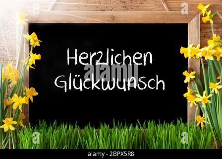 Lavagna con testo tedesco Herzlichen Glueckwunsch significa Congratulazioni. Fiori di primavera assolati Nacissus o Daffodil con erba. Rustico in legno invecchiato Ba Foto Stock