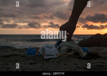 Uomo habd raccogliere rifiuti medici, maschera e guanti di plastica rifiuti rifiuti rifiuti sulla spiaggia del mare tramonto, coronavirus covid malattia di inquinamento Foto Stock