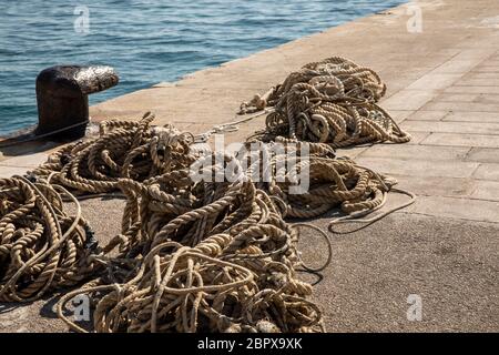 Funi ormeggiando su un vecchio tollard di metallo e il mare azzurro limpido sullo sfondo. Molte corde di diverse dimensioni che si stese a terra al porto Foto Stock