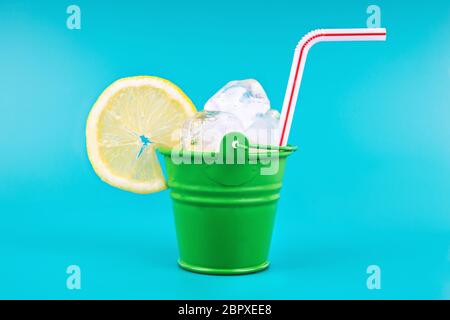 Primo piano di un secchio di acciaio verde con cubetti di ghiaccio, fetta di limone e una paglia su uno sfondo blu intenso. Secchio completo di un cocktail rinfrescante. Foto Stock