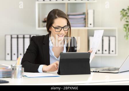Felice lavoratore di ufficio lavora confrontando i documenti su carta online su un tablet Foto Stock