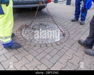 Tre lavoratori inriconoscibili dei servizi pubblici all'apertura di una pesante doppia tombatura con gru e catena in acciaio e agganciati in un pedone Foto Stock