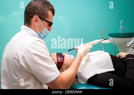 Dentista che usa una lampada ultravioletta sbiancante sui denti. La giovane paziente della clinica dentale si trova in occhiali uv Foto Stock