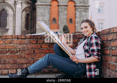 Bella ragazza in una camicia plaid disegna vecchi edifici mentre si siede vicino a un muro di mattoni. La ragazza trascorre il tempo libero disegnando immagini su una strada cittadina. Foto Stock