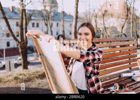 Un'artista giovane positiva disegna un'immagine mentre si siede su una panchina. Un artista sorridente tiene in mano una tavoletta di legno per la pittura e un pennello Foto Stock