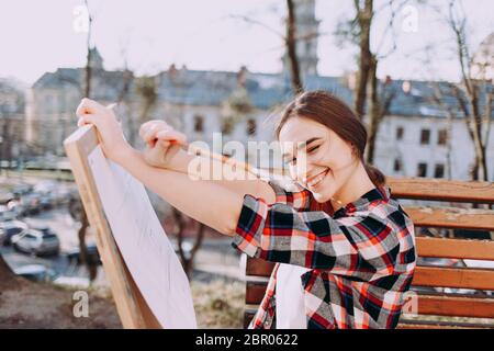 Un'artista giovane positiva disegna un'immagine mentre si siede su una panchina. Un artista sorridente tiene in mano una tavoletta di legno per la pittura e un pennello Foto Stock