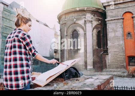 Donna sta dipingendo direttamente sulla strada della città sullo sfondo di una vecchia architettura. Giovane pittore in opera Foto Stock