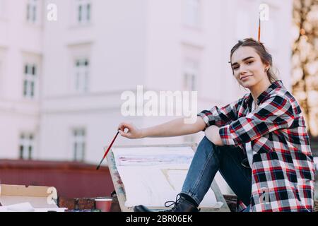 Artista donna dipinge una foto mentre si siede su un muro di mattoni su uno sfondo di vecchia architettura. La ragazza trascorre il tempo libero disegnando immagini su una strada cittadina Foto Stock