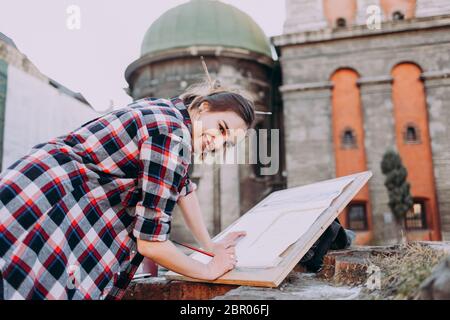 Bella giovane artista donna dipinge l'architettura vecchia in città. La ragazza trascorre il tempo libero disegnando immagini su una strada cittadina. Sorridente artista femminile Foto Stock