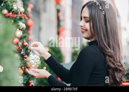 Bella ragazza giovane pende palle su un albero di Natale vicino al negozio. Cute sorridente ragazza decora un albero di Natale Foto Stock