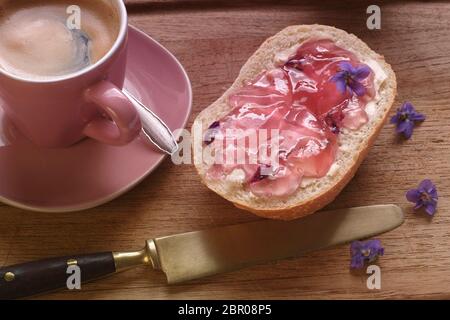 Un rotolo di pane con gelatina viola accanto a una tazza di caffè. Sui panini accanto alle violette fresche sono per decorazione. La scena è fotografata in paesaggio Foto Stock