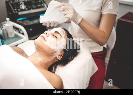 Il cosmetologo lava la maschera dal viso del paziente. Rimuovere una maschera idratante dal viso Foto Stock