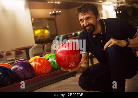 Il bowler sorridente sta giocando le bocce e tenendo una palla di bowling rossa in mano nella pista da bowling interna. Foto Stock