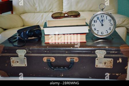 Mucchio di libri e sveglia su una valigia marrone, binoculare, occhiali da sole. Foto Stock