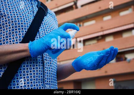 primo piano di un uomo caucasico per strada, indossando guanti blu in lattice, disinfettando le mani spruzzando un disinfettante blu da una bottiglia Foto Stock