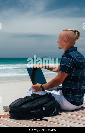 Un ragazzo giovane guarda il mare lavorando con un computer portatile, seduto sulla riva di una spiaggia di sabbia. Uomo d'affari in un viaggio di lavoro remoto. Blogger, fr Foto Stock