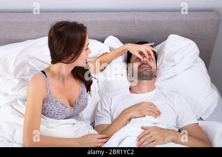 Disturbato giovane donna tenendo il marito del naso per farlo smettere di russare sul letto Foto Stock