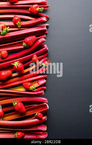 Fragole e steli di rabarbaro allineati su un tavolo nero, sopra la vista. Verdure fresche e frutta, ingredienti per una torta Foto Stock