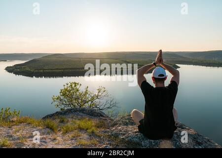 Escursionista maschile facendo yoga sulla roccia vicino ampio fiume. Un uomo sta pregando Dio sulla montagna Foto Stock