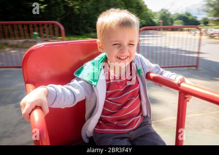 Poco carino ragazzo caucasico spinning round di posti a sedere su una giostra sedia in metallo in un parco giochi in estate Foto Stock