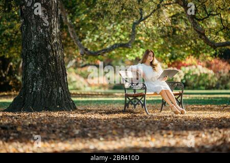 Bella donna asiatica in abito bianco sedersi sotto la scrittura albero e pensare nel parco con foglie d'autunno. Foto Stock