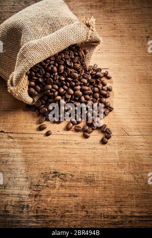 Piena arrosto di miscele di caffè in grani fuoriuscita da un piccolo sacchetto di tela su legno rustico con spazio di copia in formato verticale Foto Stock