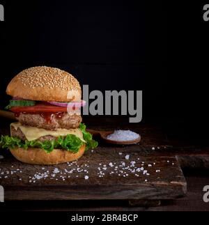 Cheeseburger in un panino con semi di sesamo, nel mezzo di verdure fresche e costolette di carne, sfondo nero Foto Stock