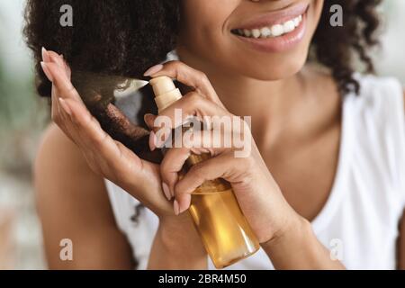 Trattamento con estremità separate. Donna nera sorridente spruzzando olio essenziale sui capelli ricci Foto Stock