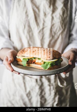 Donna in grembiule di lino con panino di pesce fresco fritto con salsa tartare, limone e rucola, fuoco selettivo. Idee per la colazione salutari e facili Foto Stock