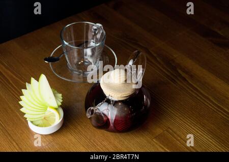 Un bicchiere di infuso di tè e un bicchiere con il limone in una teiera di vetro collocato su un tavolo marrone Foto Stock