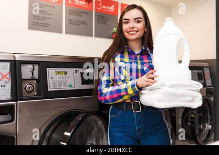 La ragazza soddisfatta nella lavanderia tiene una pila di asciugamani bianchi e detersivo della biancheria Foto Stock