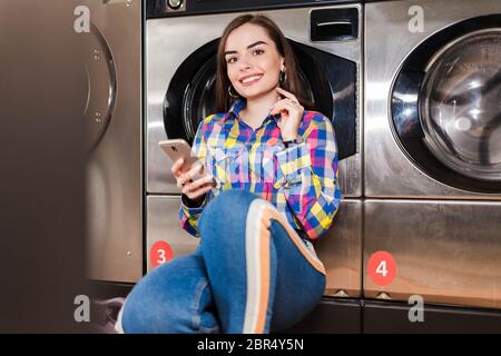 Bella ragazza con smartphone in mano ascolta la musica appoggiata su una lavatrice in lavanderia pubblica Foto Stock