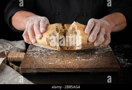 Baker in uniforme nera si è rotto in metà tutta una pagnotta cotta di farina di grano bianco pane Foto Stock