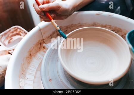 Vasaio professionale lavora su lastre di verniciatura in officina. Donna dipinge una piastra in ceramica con un pennello e vernice blu Foto Stock