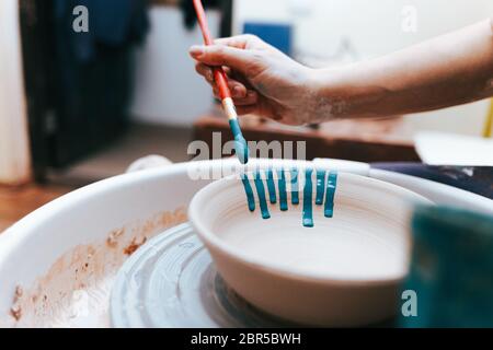 Vasaio professionale lavora su lastre di verniciatura in officina. Donna dipinge una piastra in ceramica con un pennello e vernice blu Foto Stock