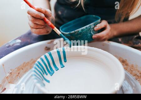 Vasaio professionale lavora su lastre di verniciatura in officina. Donna dipinge una piastra in ceramica con un pennello e vernice blu Foto Stock