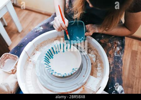 Vasaio professionale lavora su lastre di verniciatura in officina. Donna dipinge una piastra in ceramica con un pennello e vernice blu Foto Stock