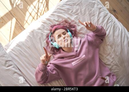 Felice ragazza rosa capelli indossare cuffie sdraiati a letto ascoltando musica canto. Foto Stock