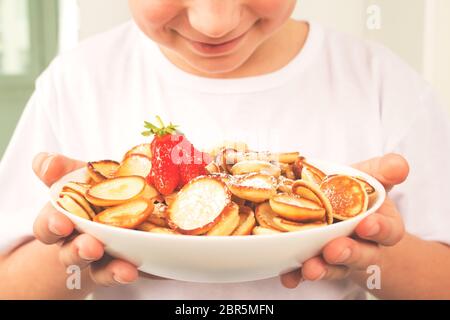 Happy boy con ciotola bianca con mini pancake sani alla moda, cereali e fragole. Nessuna immagine del volto, concetto di famiglia. Sana colazione vegana senza Foto Stock