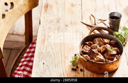 Pentola di Rame del ricco gustoso cervo Spezzatino di cervo condita con erbe aromatiche servita su una tavola in legno rustico con condimento fresco, bicchiere di vino rosso e palchi Foto Stock