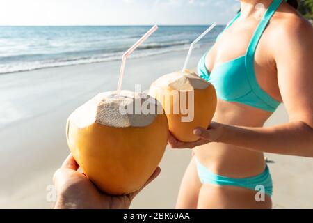 Close-up del giovane azienda noce di cocco con cannuccia in piedi presso la spiaggia vicino al mare Foto Stock