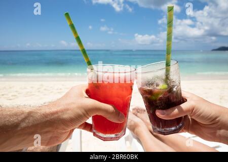 Close-up del giovane la mano il tintinnio dei bicchieri di succo di frutta con cannuccia sulla spiaggia Foto Stock