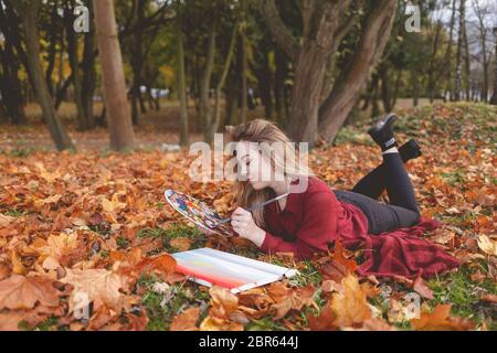 Giovane artista donna si sta rilassando nel parco per il suo hobby preferito. La ragazza disegna un'immagine, tenendo una tavolozza in mano Foto Stock