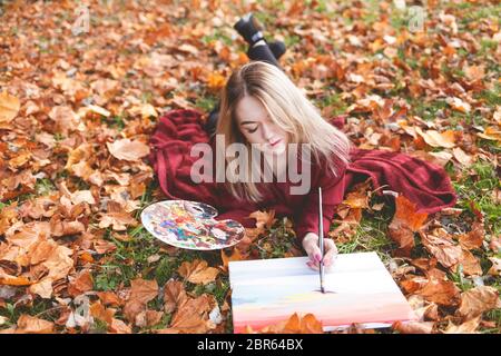 Giovane artista donna si sta rilassando nel parco per il suo hobby preferito. La ragazza disegna un'immagine, tenendo una tavolozza in mano Foto Stock