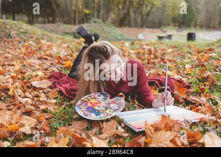 Giovane artista si trova nel parco autunnale su una plaid e dipinge un quadro. La ragazza disegna una foto nel parco Foto Stock