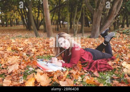 Giovane artista si trova nel parco autunnale su una plaid e dipinge un quadro. La ragazza disegna una foto nel parco Foto Stock