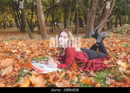 Giovane artista si trova nel parco autunnale su una plaid e dipinge un quadro. La ragazza disegna una foto nel parco Foto Stock