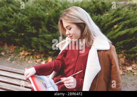 Bionda donna disegna una foto su una panchina del parco. Artista impegnato nel suo hobby preferito di disegno. Foto Stock